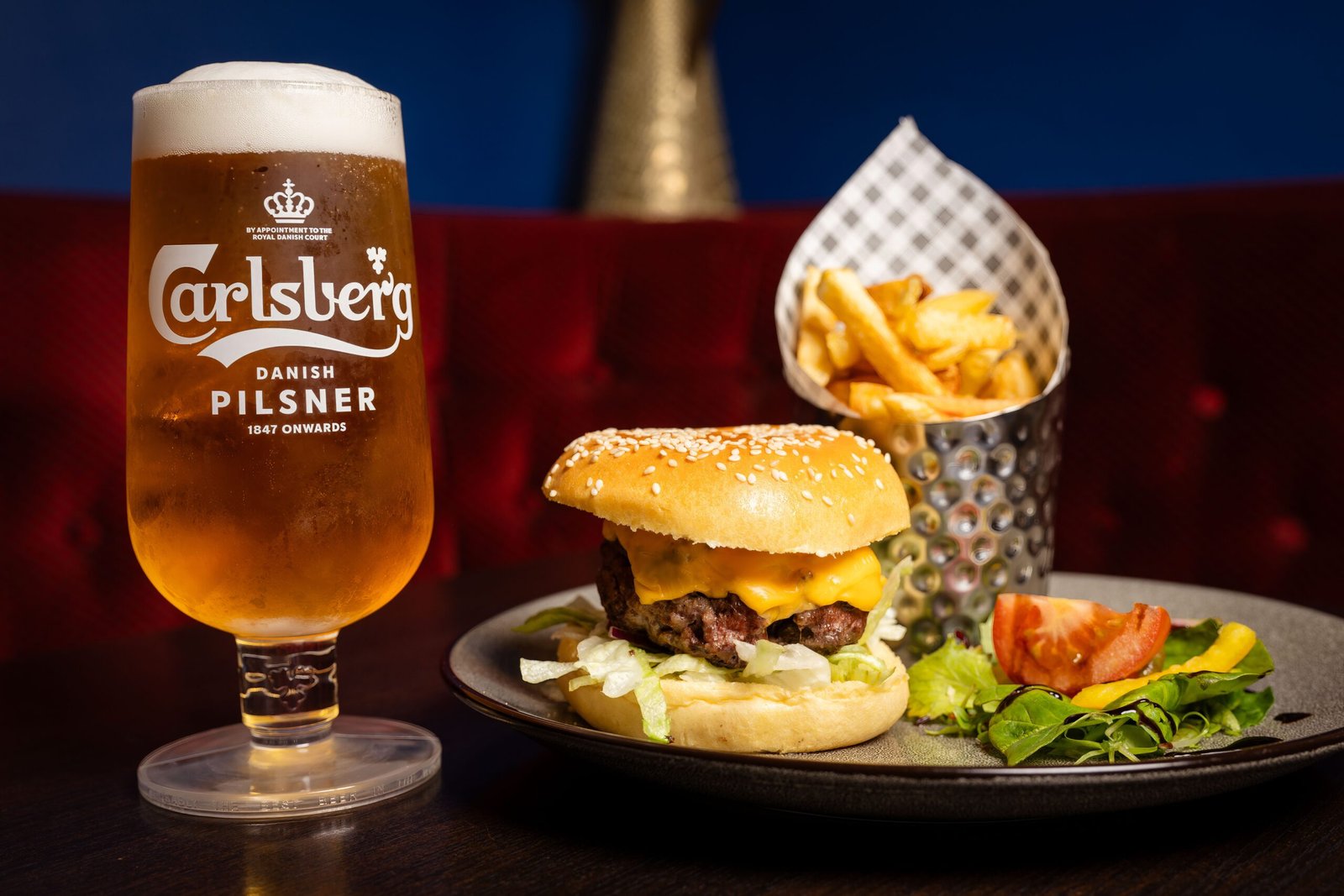 A pint of Carlsberg beer is served alongside a cheeseburger with lettuce, tomato, and a side of fries on a plate, set against a cozy pub backdrop.