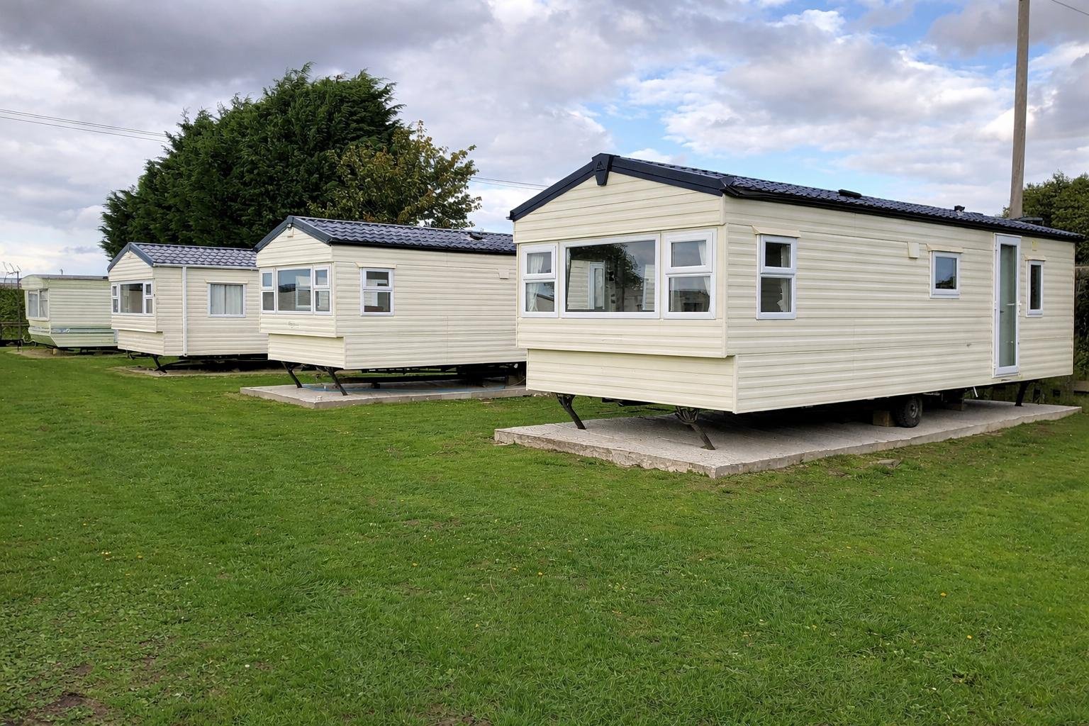 Caravan chalets lined up on a grassy area at Withamside Country Park, surrounded by trees and under a partly cloudy sky. The setting offers a peaceful atmosphere, ideal for a countryside getaway.